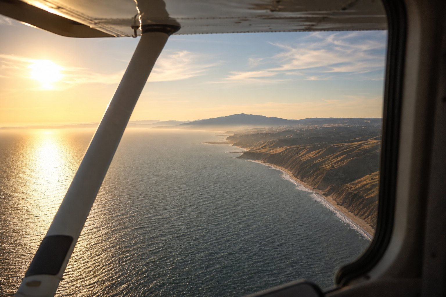 A coastline at golden hour seen past the wing strut from the passenger seat of a small propeller aircraft