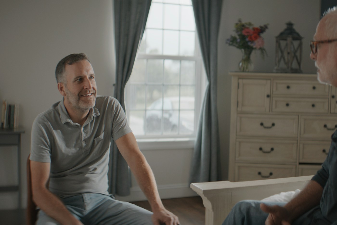 Two men talking across a sunlit room, one older, one listening